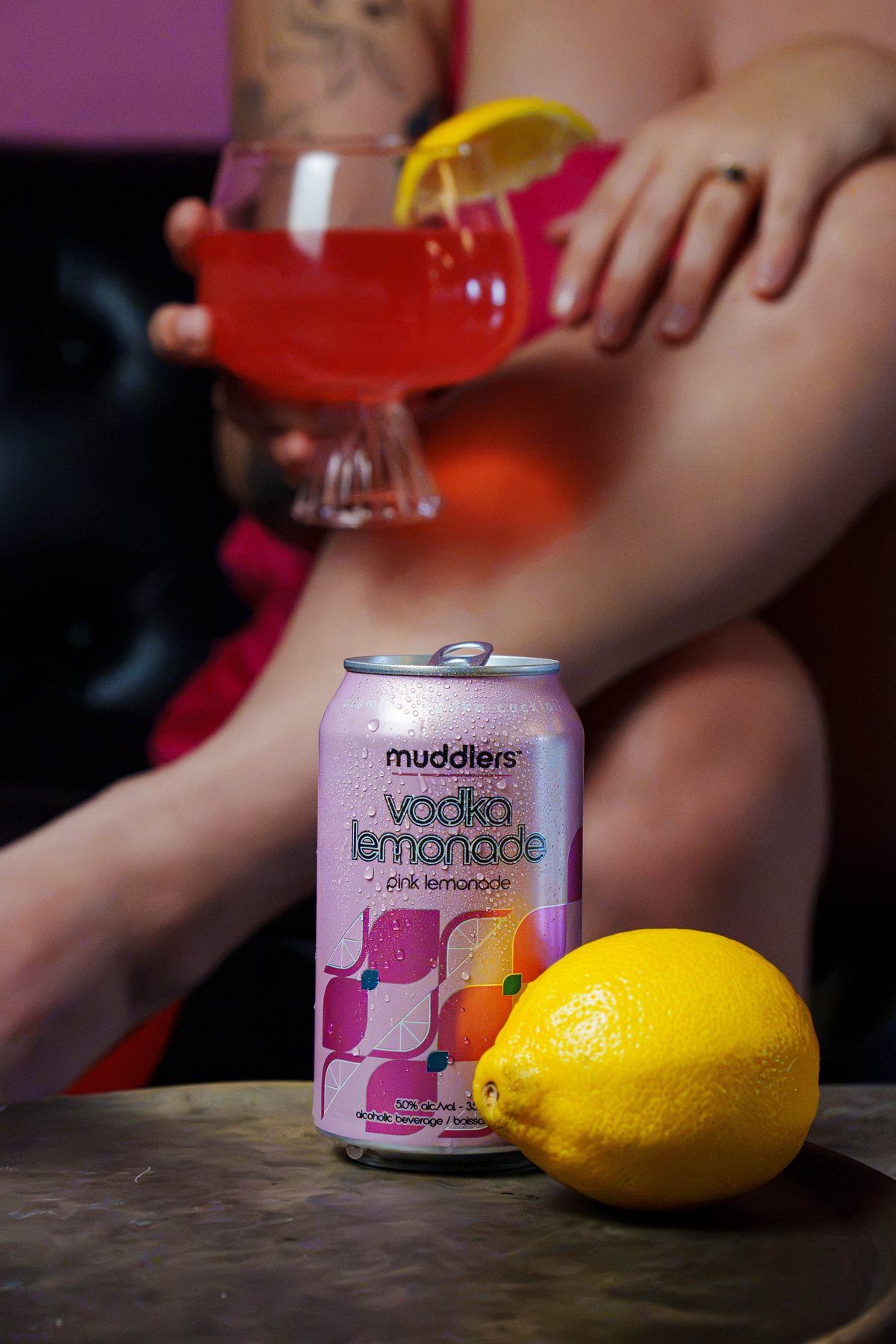 A can of Muddlers Vodka Pink Lemonade beside a lemon, with a glass of it held in the background